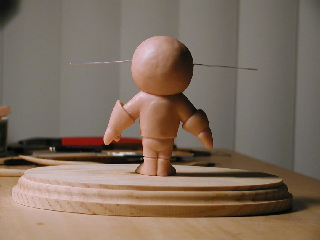 A small figurine with a large head, no facial features, and tiny limbs, standing on a wooden base with a spinning top mechanism, with a thin wire connected horizontally through its head.