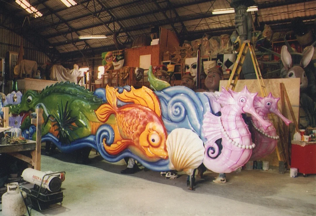 Colorful horse and sea creature parade float in a workshop with masks and sculptures in the background.