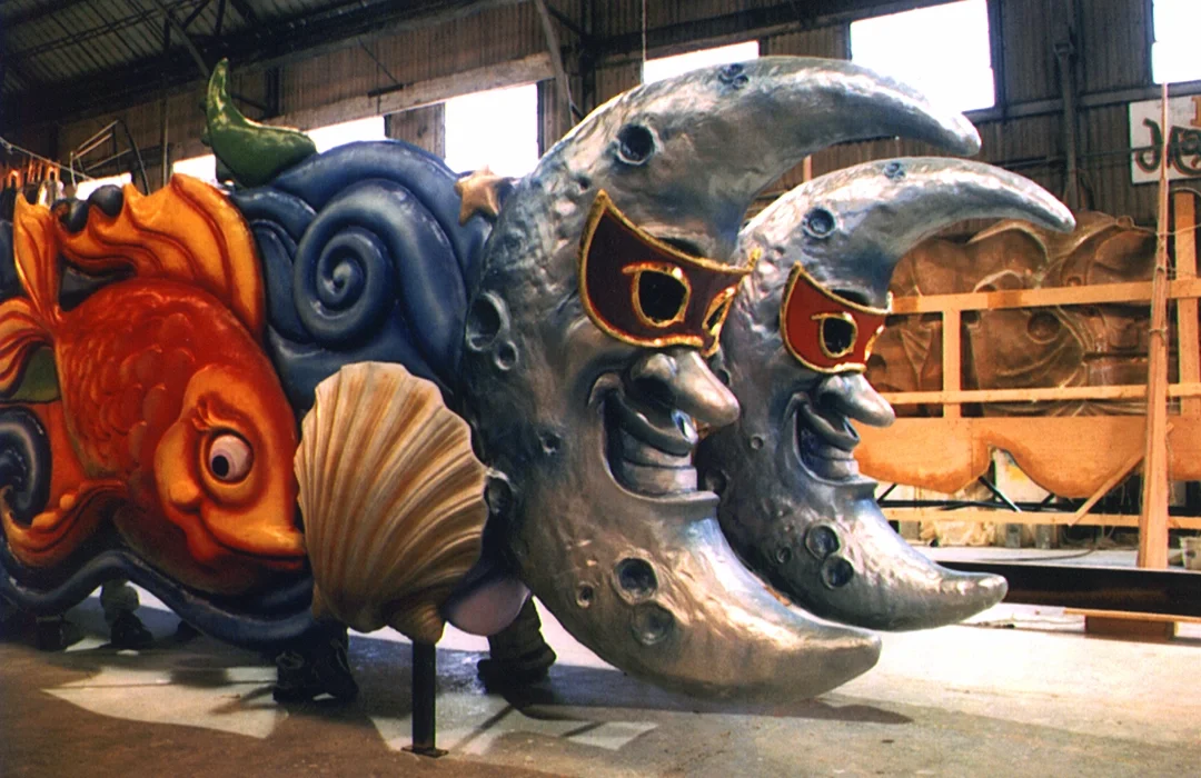 Colorful parade float with a large fish, seashell, and moon face with sunglasses inside a warehouse.