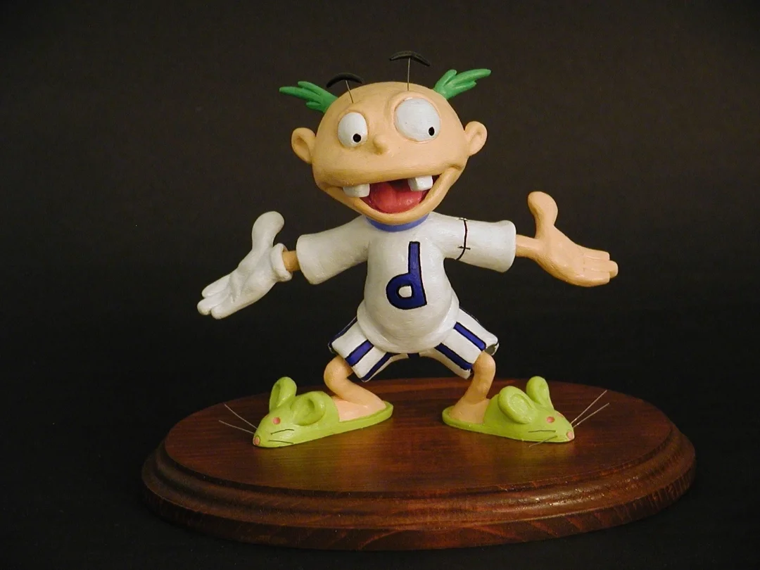 A cartoonish figurine of a boy with green hair, big eyes, and buck teeth, standing on a wooden base with green mice shoes, wearing a white t-shirt with the letter 'd' and striped shorts, smiling with arms outstretched against a black background.