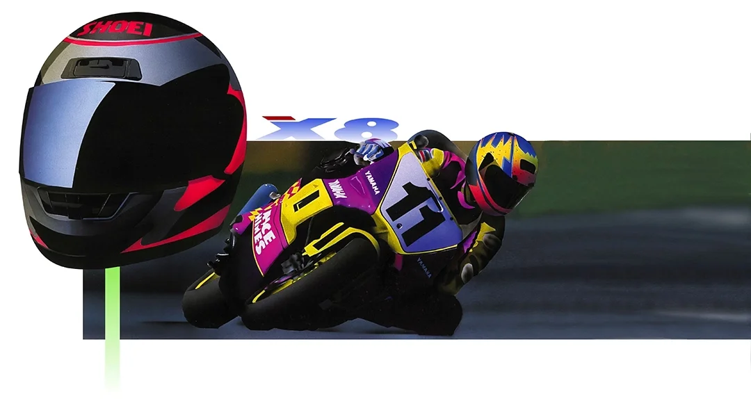 Motorcycle racer in yellow and purple gear leaning into a turn on a racetrack, with a black helmet featuring colorful accents and a large helmet with red, black, and gray design in the background.