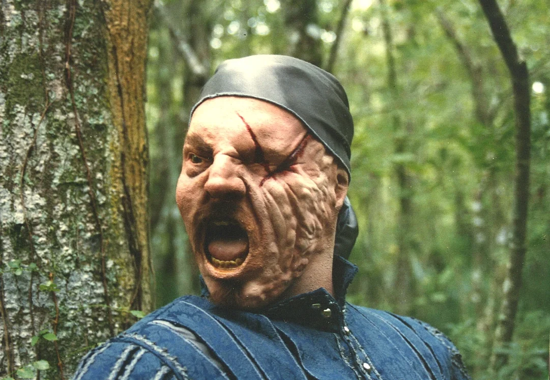 A person wearing a bandana makes a face with a horror-movie mask with a large scar and bulging skin, in a wooded outdoor setting.