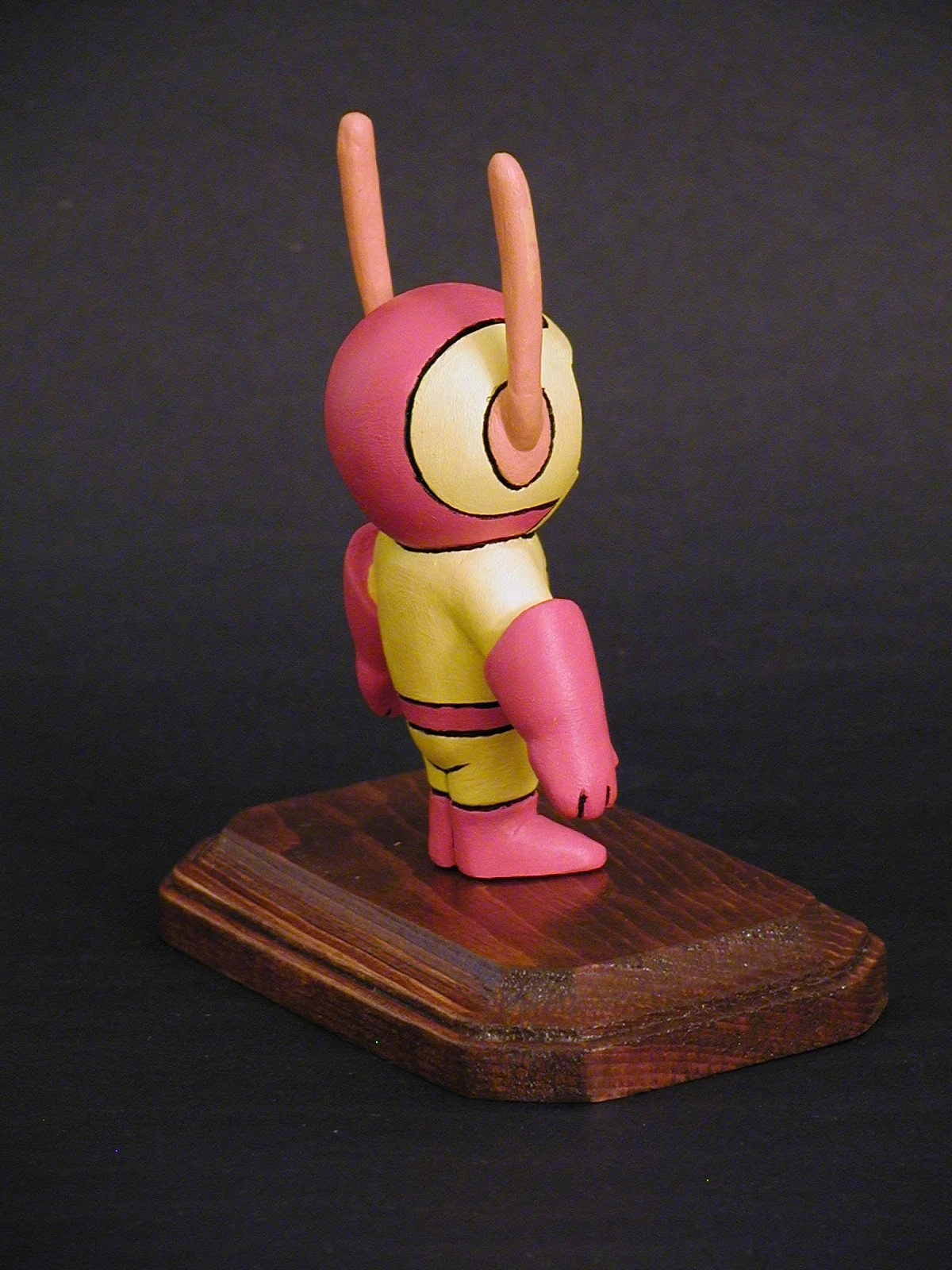 A colorful cartoon-style figure with a round head, two antennae, pink and yellow body, and pink boots, standing on a wooden platform against a dark background.