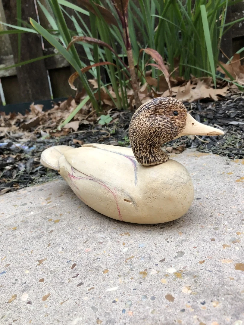 Wooden duck figurine with a carved and painted duck head, placed on concrete outdoors with plants and leaves in the background.