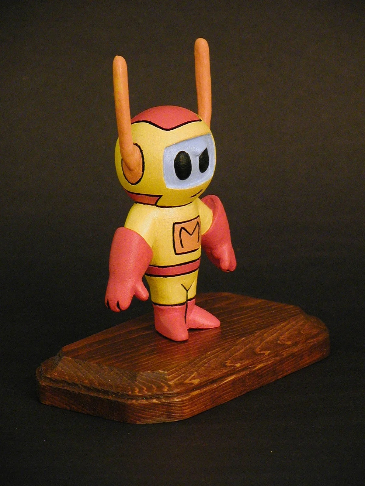 Colorful cartoon-like robot figure with antennae, standing on a wooden base, against a dark background.