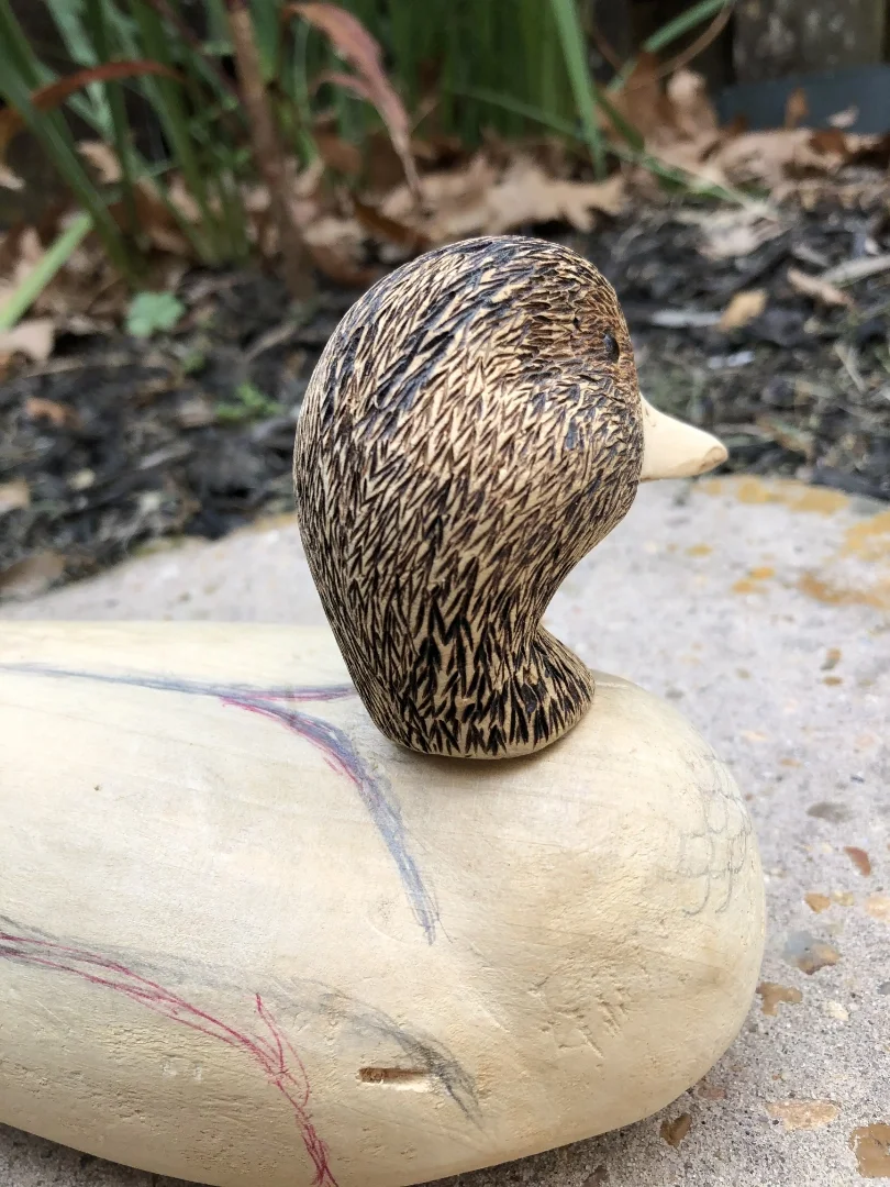 Wooden sculpture of a duck, featuring a textured, detailed head and beak, placed on a light-colored, rounded wooden base outdoors.