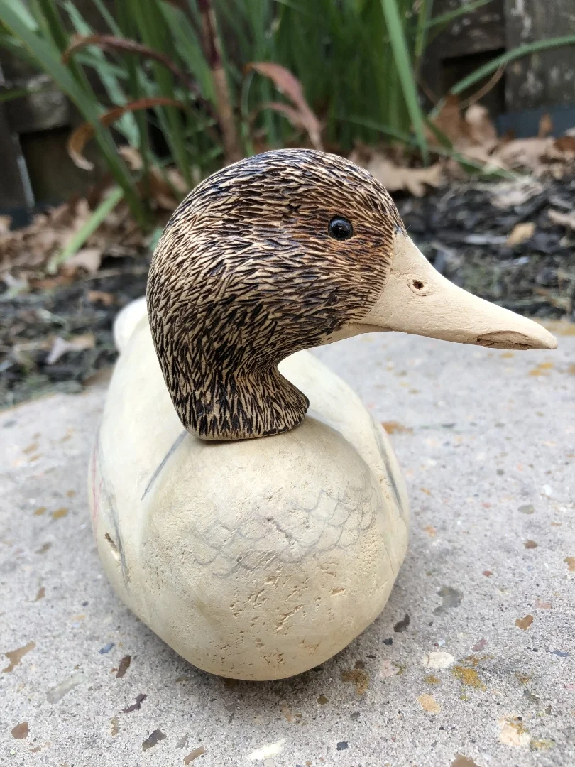 A decorative duck figurine with a carved wooden head and a stone body, placed outdoors on a concrete surface with plants and leaves in the background.