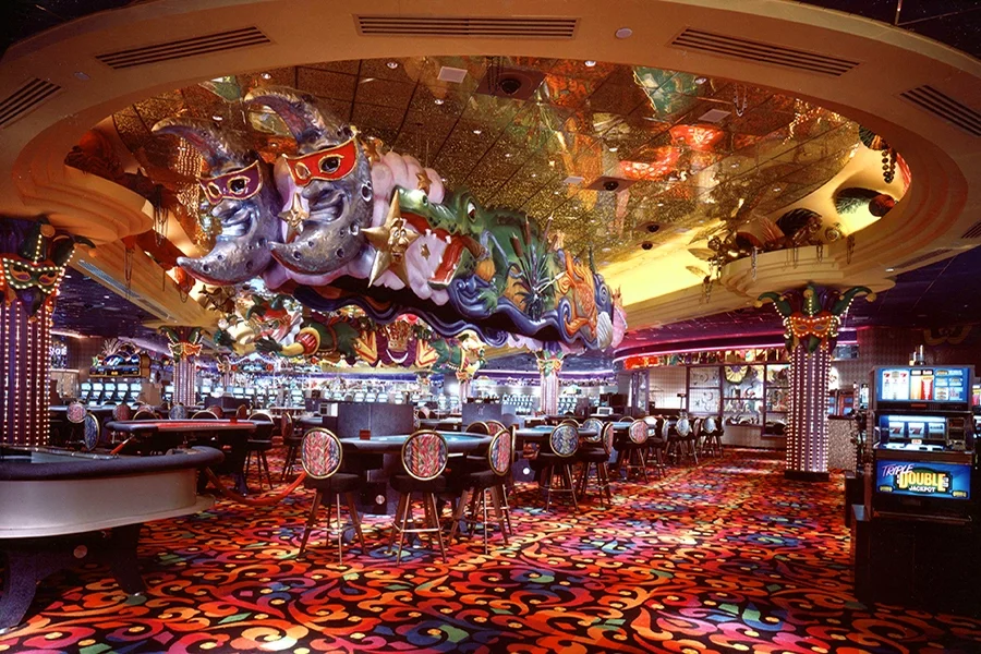 Interior of a brightly lit casino with colorful carpet, slot machines, and a bar area. Floating whimsical sculptures of moons, stars, and other shapes hang from the ceiling. The space is decorated with shiny, reflective surfaces and vibrant colors.