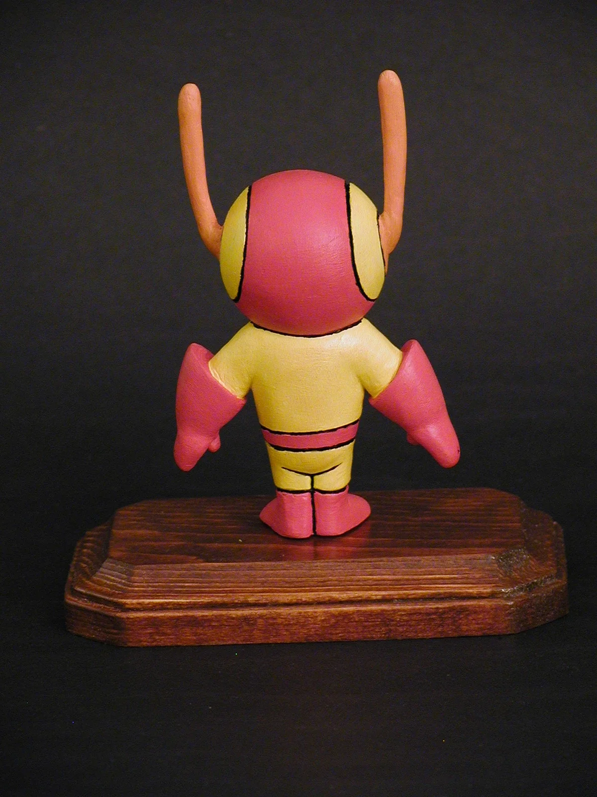 A colorful cartoon-style figurine of a character with a large round head, antennae, and pink and yellow clothes, facing away, mounted on a wooden base.