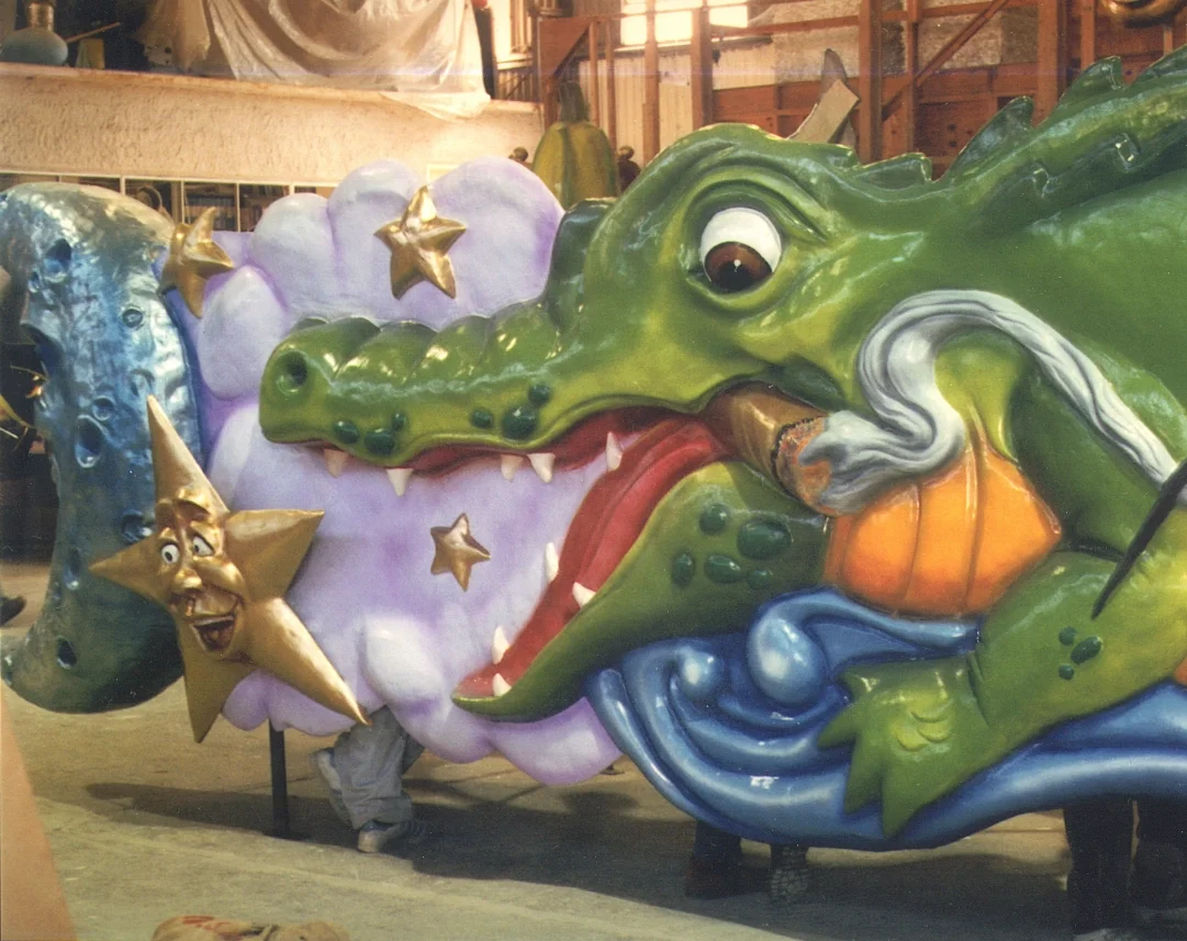 Large colorful float of a green alligator with an open mouth, wearing a captain's hat and smoking a pipe, next to a cloud with stars and a moon, all in a warehouse setting.