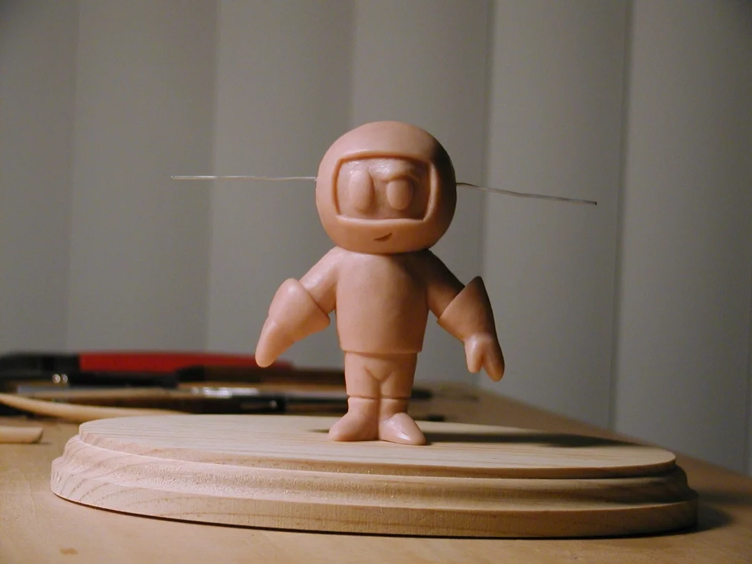 A small sculpture of an astronaut made from a material resembling clay or plastic, with exaggerated features, standing on a wooden base in a workshop setting.