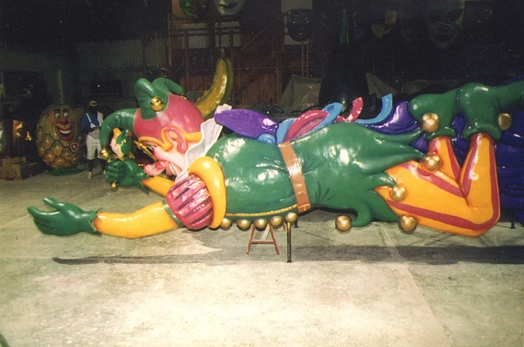 Large, colorful parade float of a jester or clown character, lying on its side, with exaggerated facial features and vibrant clothing, in a warehouse or workshop setting.
