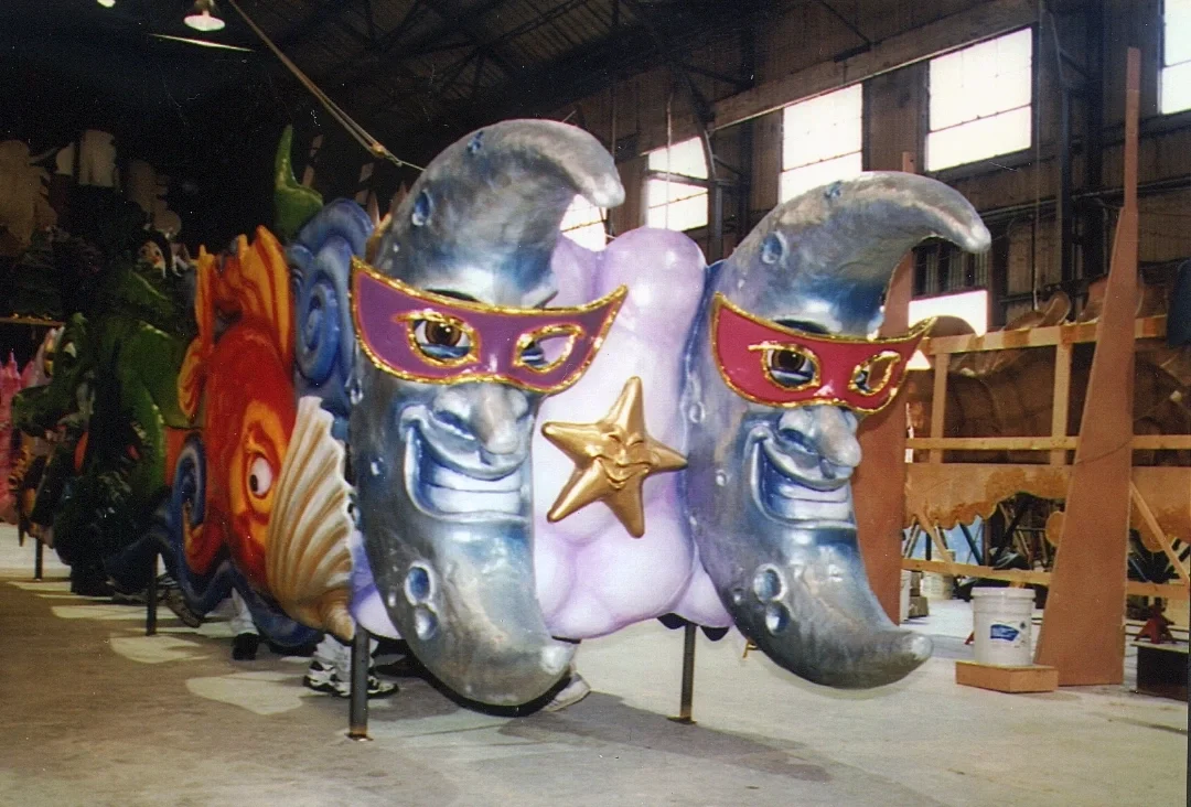 A carnival float featuring two crescent moon faces with smiling expressions, pink masks, and a gold star between them, set in an indoor warehouse.