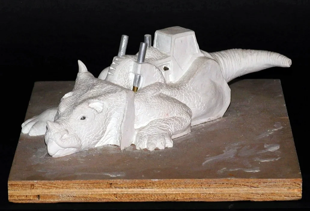 A sculpture of a large animal, resembling a crocodile, with a building on its back. The sculpture is white and appears to be made of a material like plaster, set on a wooden base.