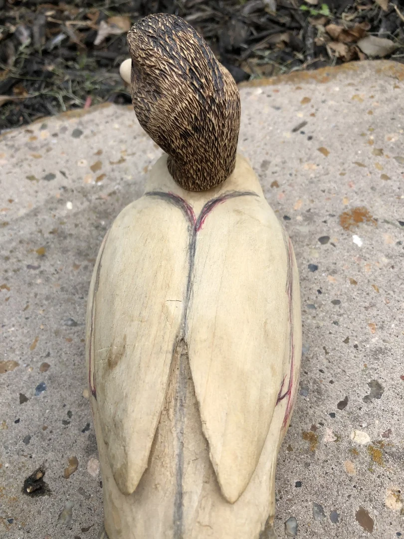 A carved wooden figure resembling an angel, with a carved head and wings painted or carved onto the body, positioned outdoors on a concrete surface.