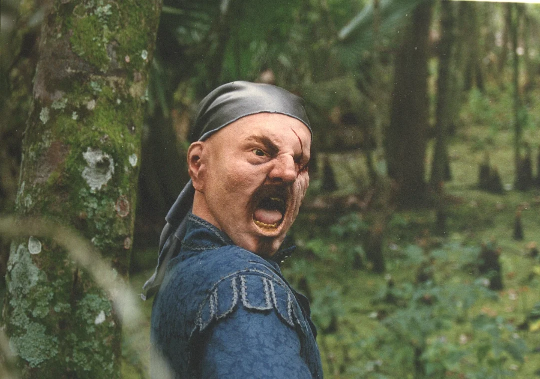 A man with a bandana on his head wearing a dark blue jacket, shouting angrily with a fierce expression, in a forest setting.