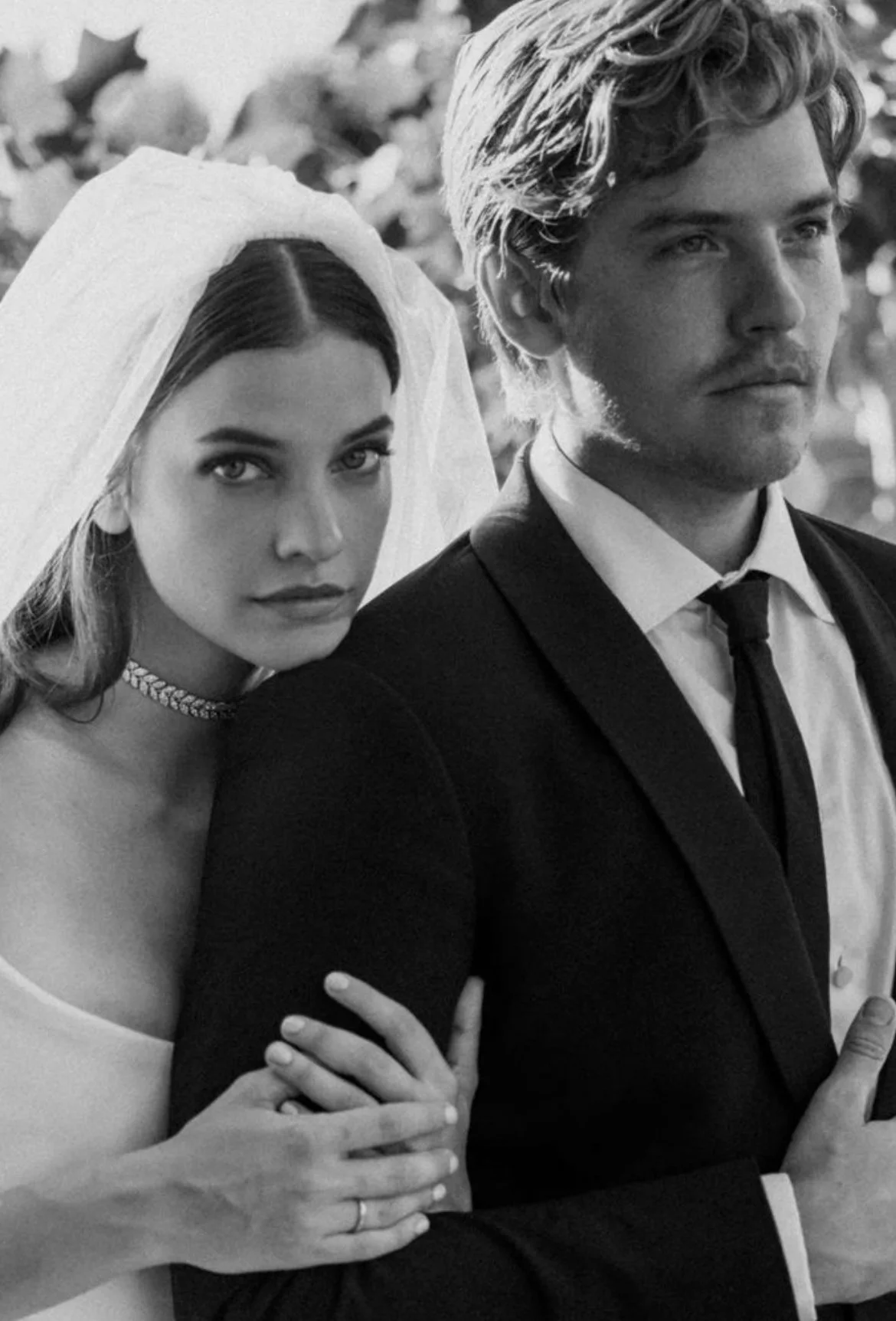 A black-and-white photo of a bride and groom. The bride has long hair, wearing a veil and a choker necklace, and is looking directly at the camera. The groom has wavy hair, wearing a suit, and is looking slightly to the side.