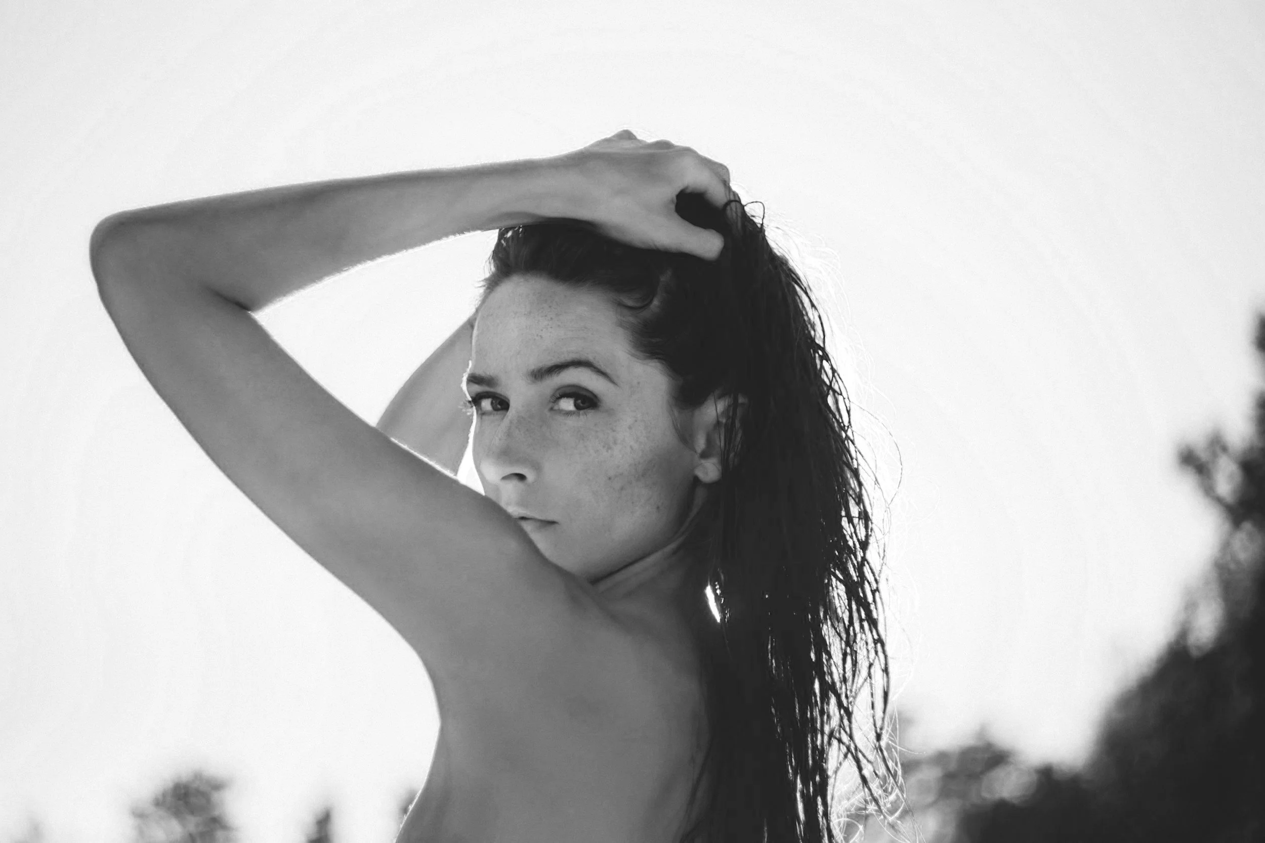A black-and-white photo of a woman with wet hair, looking over her shoulder, with one arm above her head, outdoors.