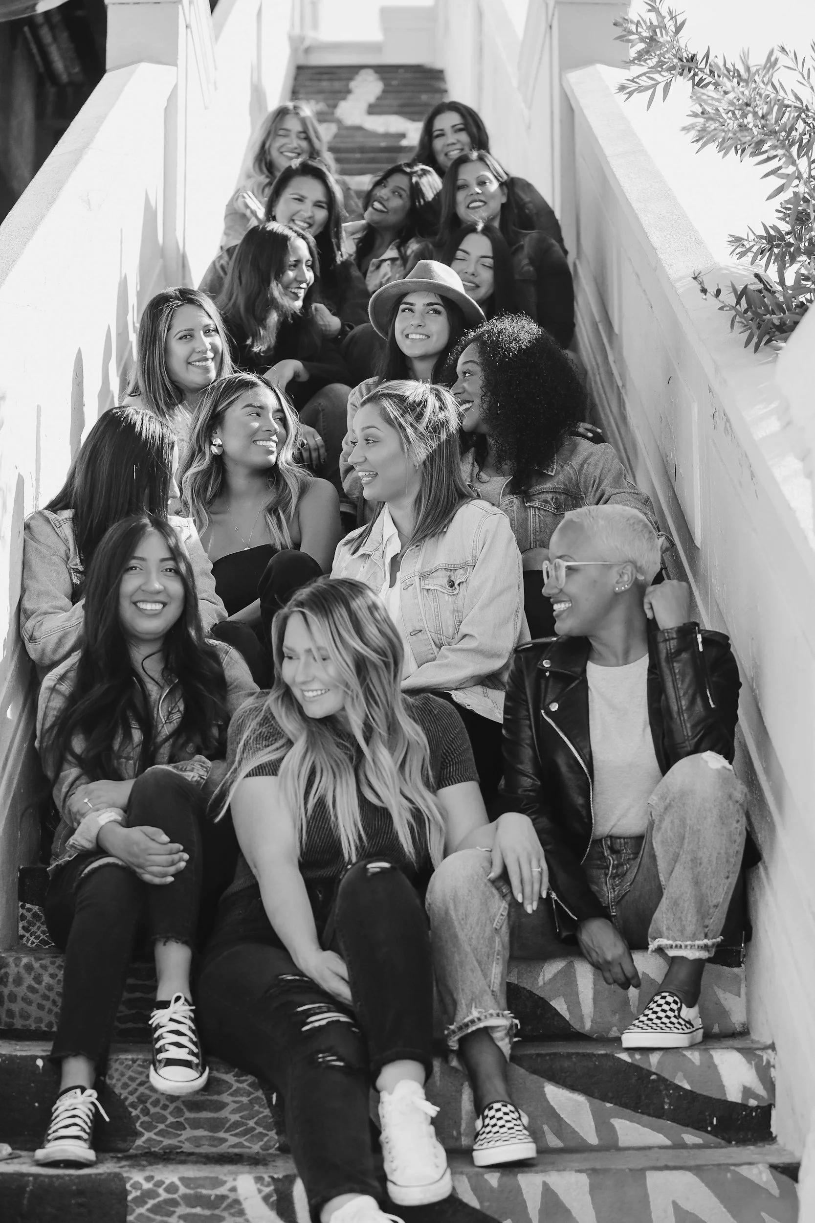 A group of women sitting on outdoor stairs, chatting and smiling, with some laughing and displaying a variety of hairstyles and casual clothing.