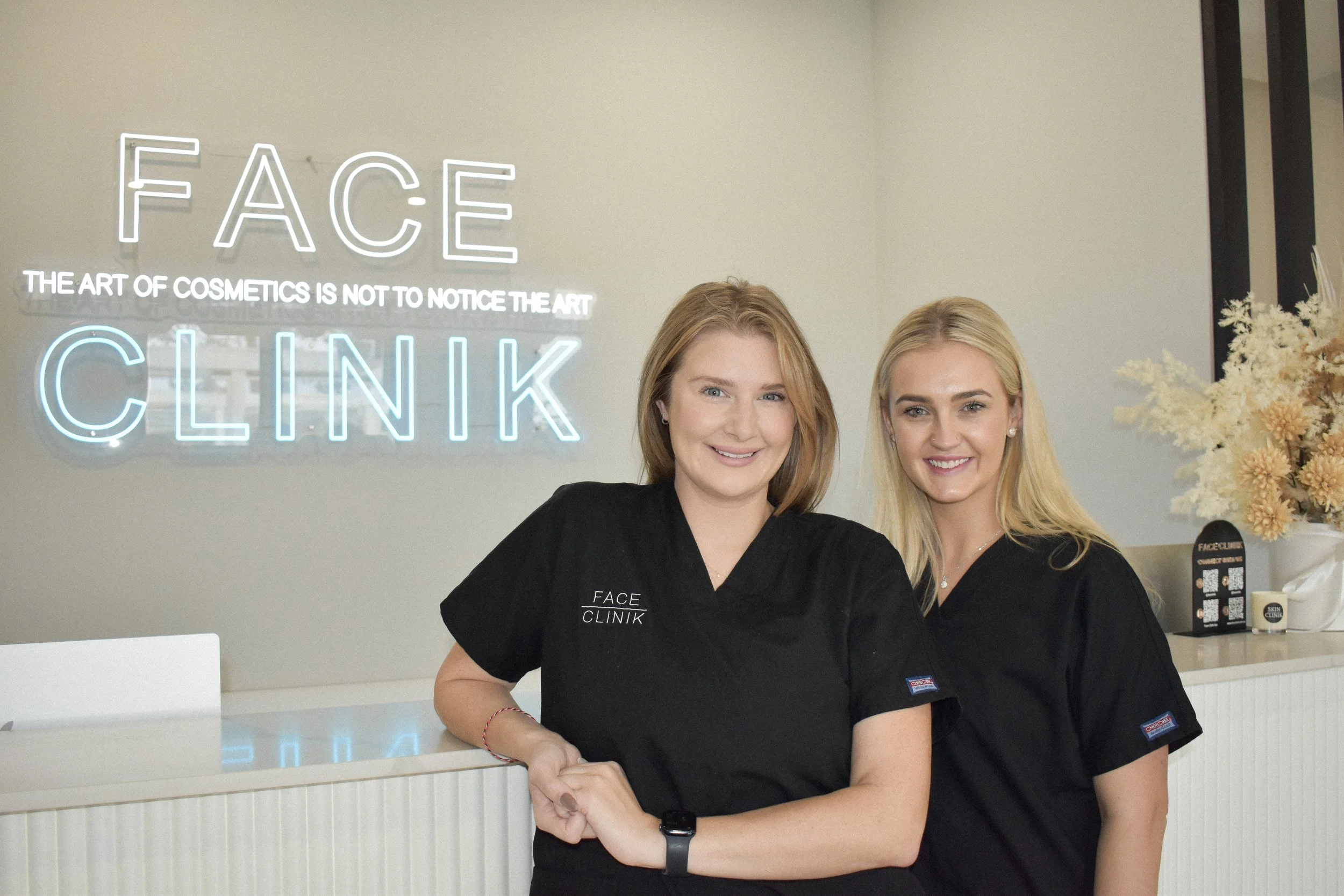 Two smiling women in black scrubs standing behind a reception desk at FACE CLINIK, a beauty or cosmetic clinic, with a neon sign on the wall behind them reading 'FACE' and 'CLINIK,' and a plant with flowers on the right side of the image.