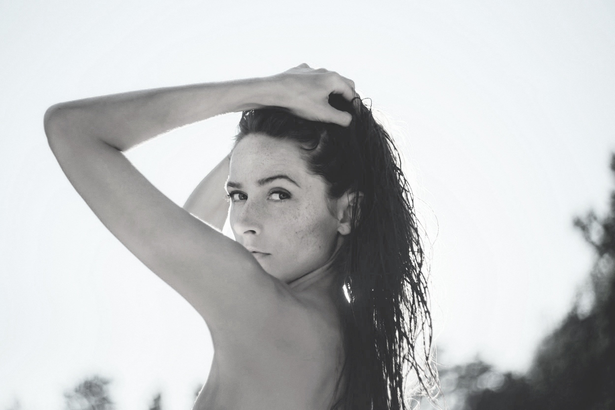 Black and white photograph of a topless woman with long, wet hair, posing outdoors with one arm raised above her head, against a clear sky, her face turned slightly to the side, with a subtle smile.