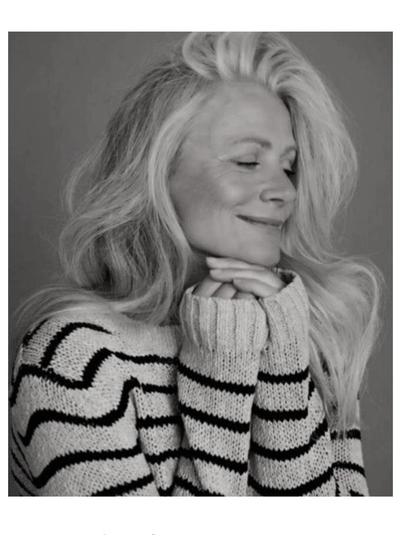 A black and white portrait of a woman with long, wavy hair, smiling softly with her eyes closed, wearing a striped sweater, resting her chin on her hands.