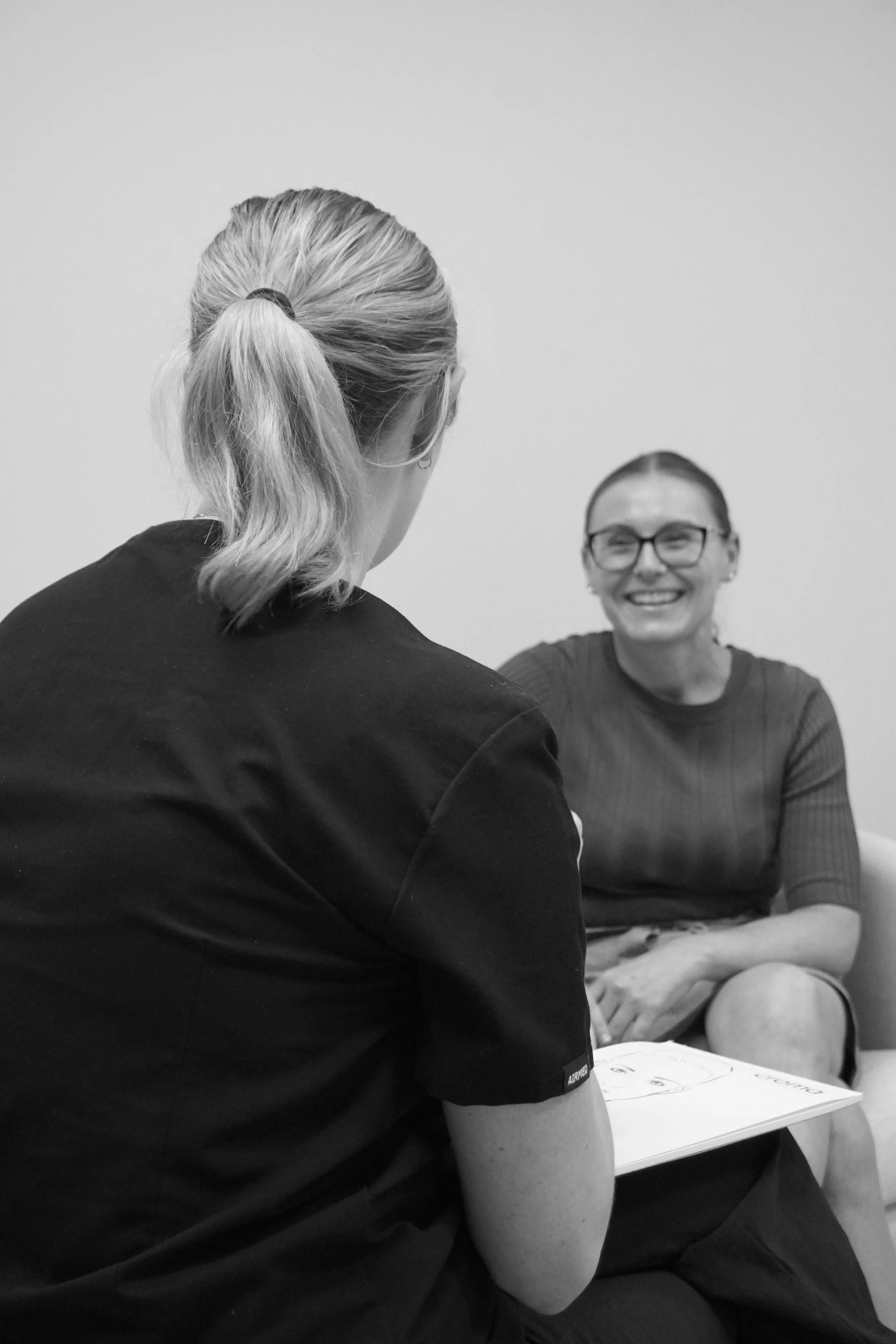 Two women engaged in a conversation, one facing away from the camera with a ponytail, the other smiling while wearing glasses, sitting on a sofa.