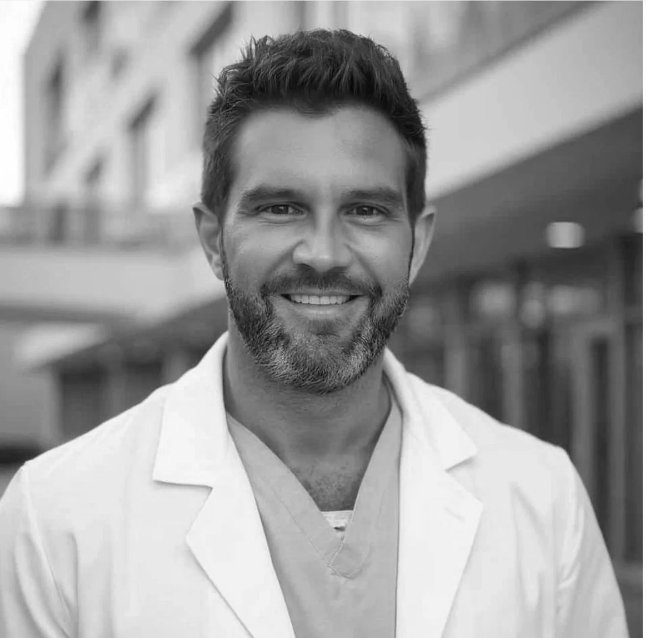 Portrait of a smiling man with dark hair and a beard, wearing medical scrubs and a white coat, outdoors with buildings in the background.