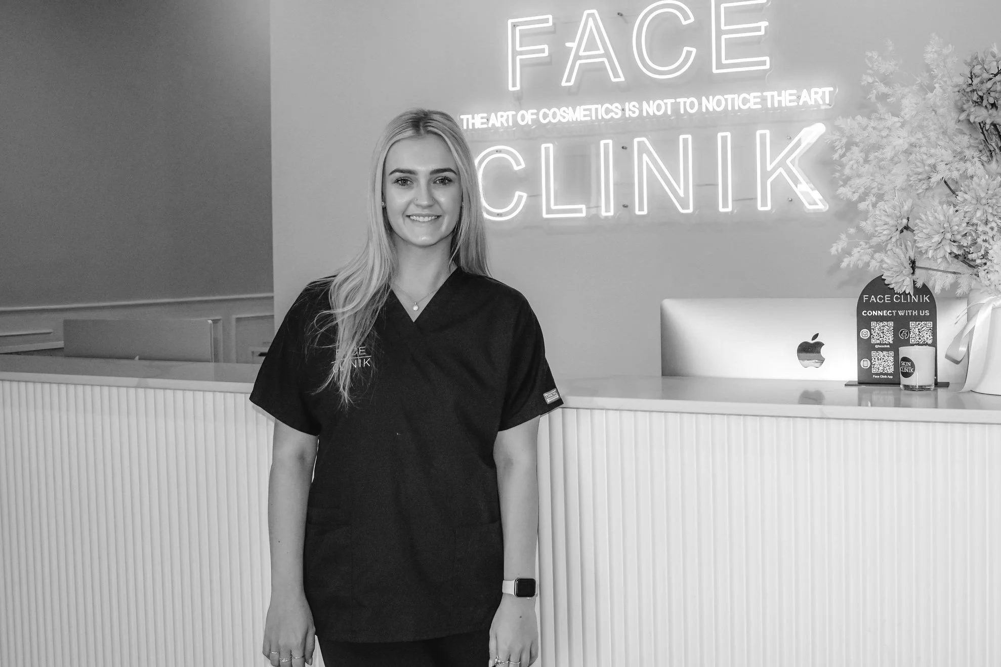 A woman with long hair smiling, wearing a black uniform, standing in front of a reception desk with a neon sign that reads 'FACE CLINIK' in a modern clinic or spa reception area.
