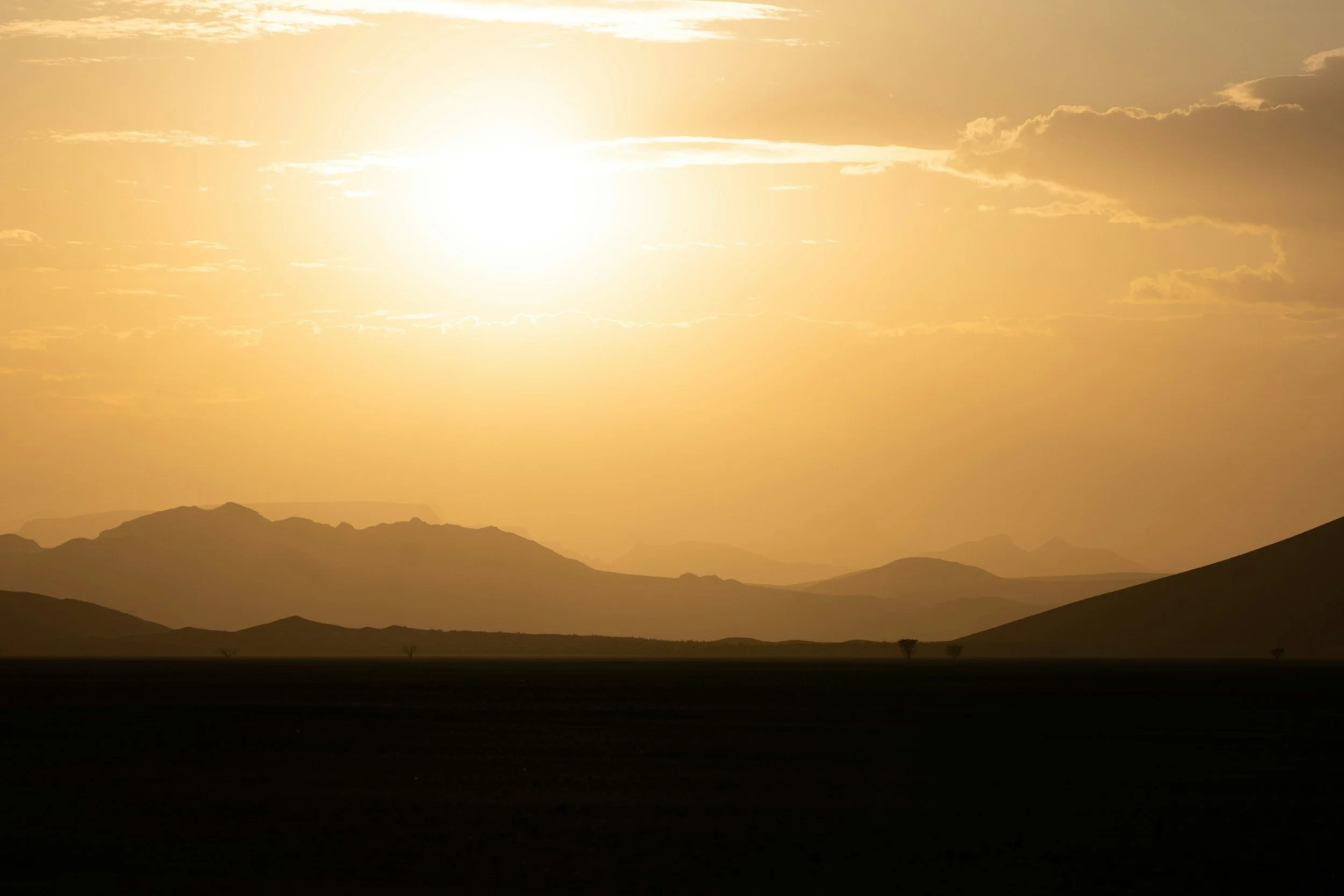 A landscape scene during sunset with mountains in the distance, a dark flat foreground, and a sky with a bright sun and scattered clouds.