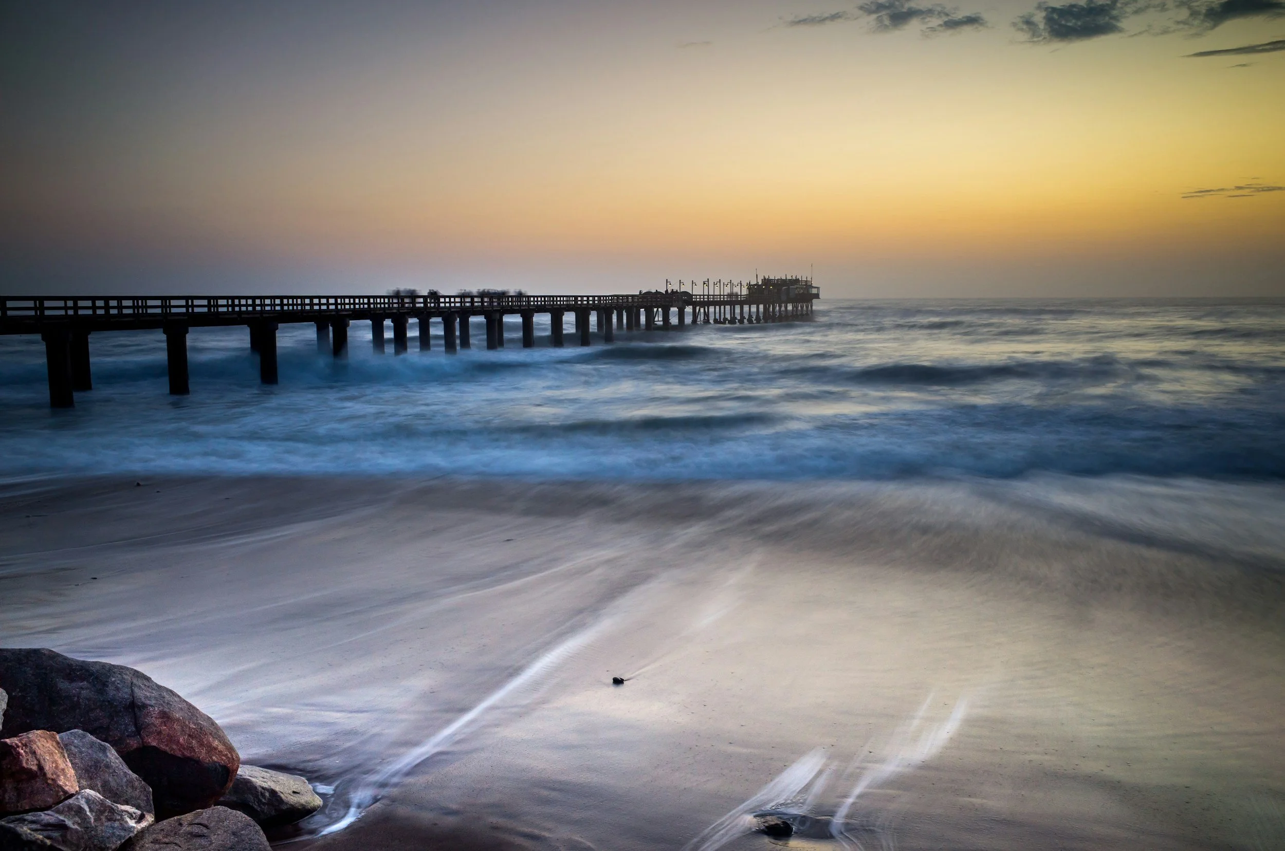 Sunset over the ocean with a wooden pier extending into the water, waves crashing on the sandy beach with rocks in the foreground.