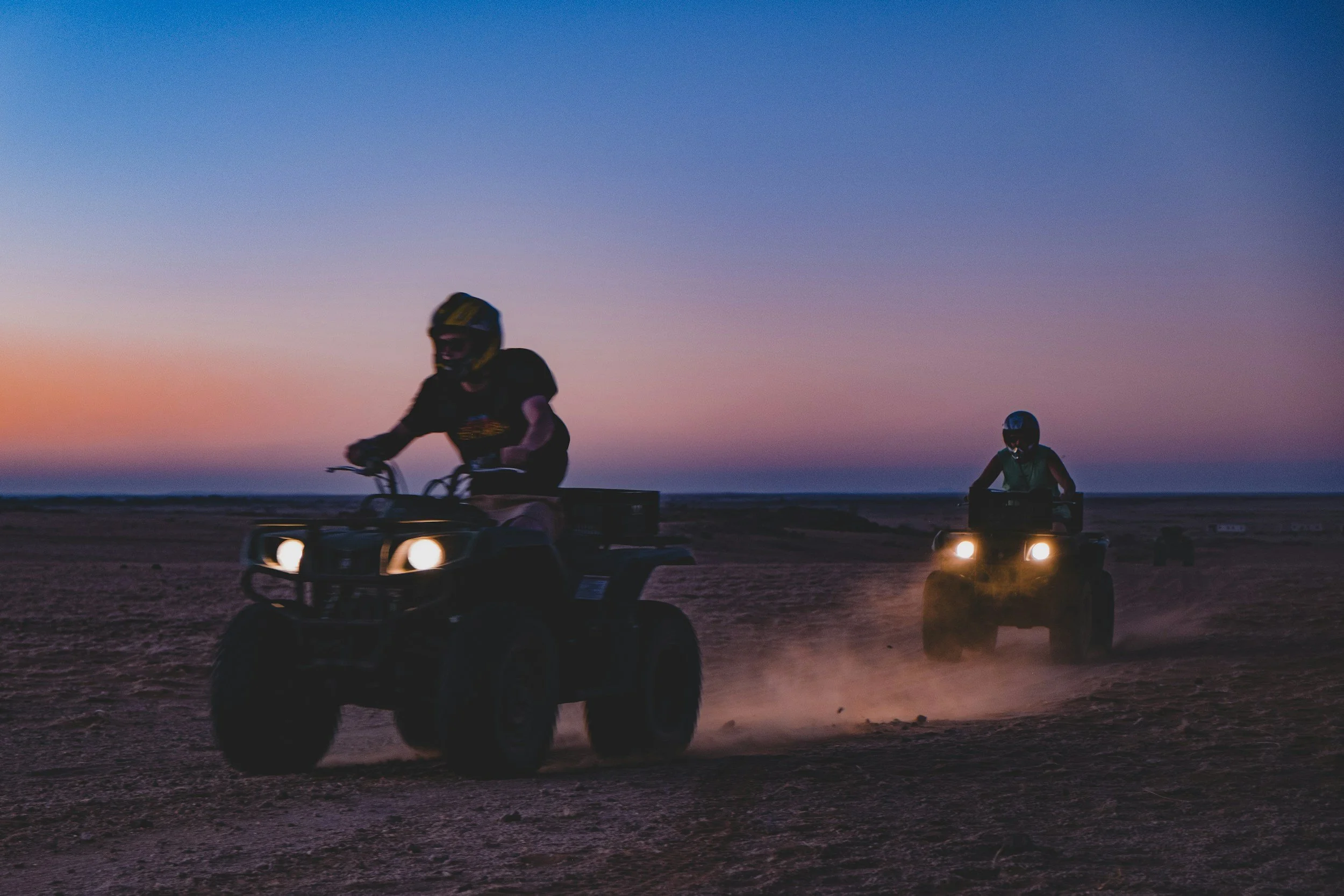 Two people riding all-terrain vehicles on a dirt path during sunset