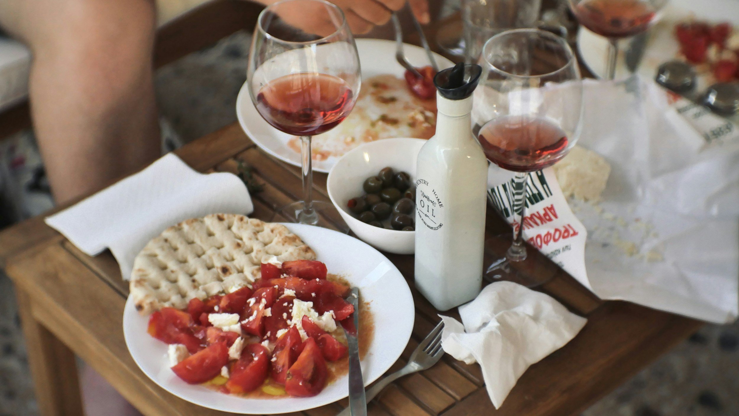 Table with sliced tomatoes, flatbread, bowl of olives, wine, and olive oil bottle.