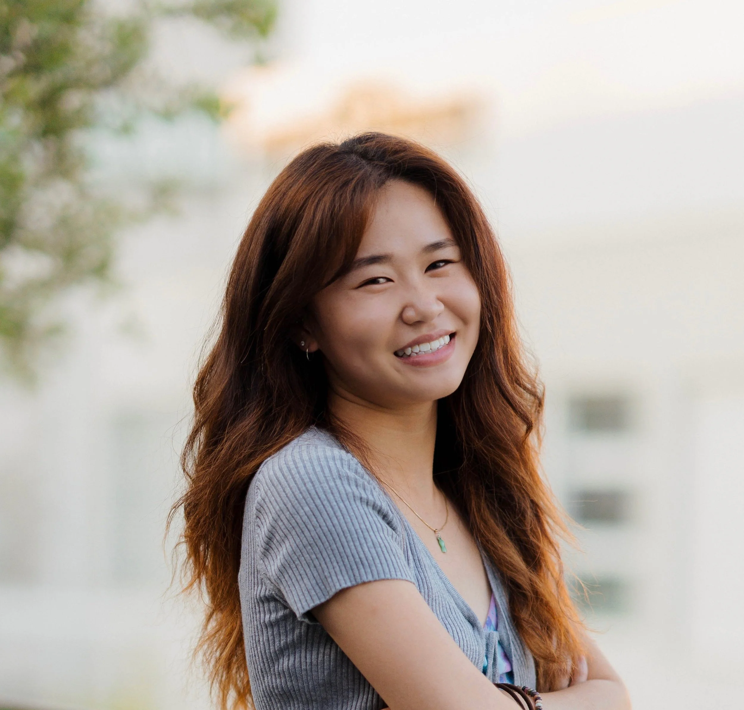 Young woman with long auburn hair, grey top, warm smile, soft outdoor background.