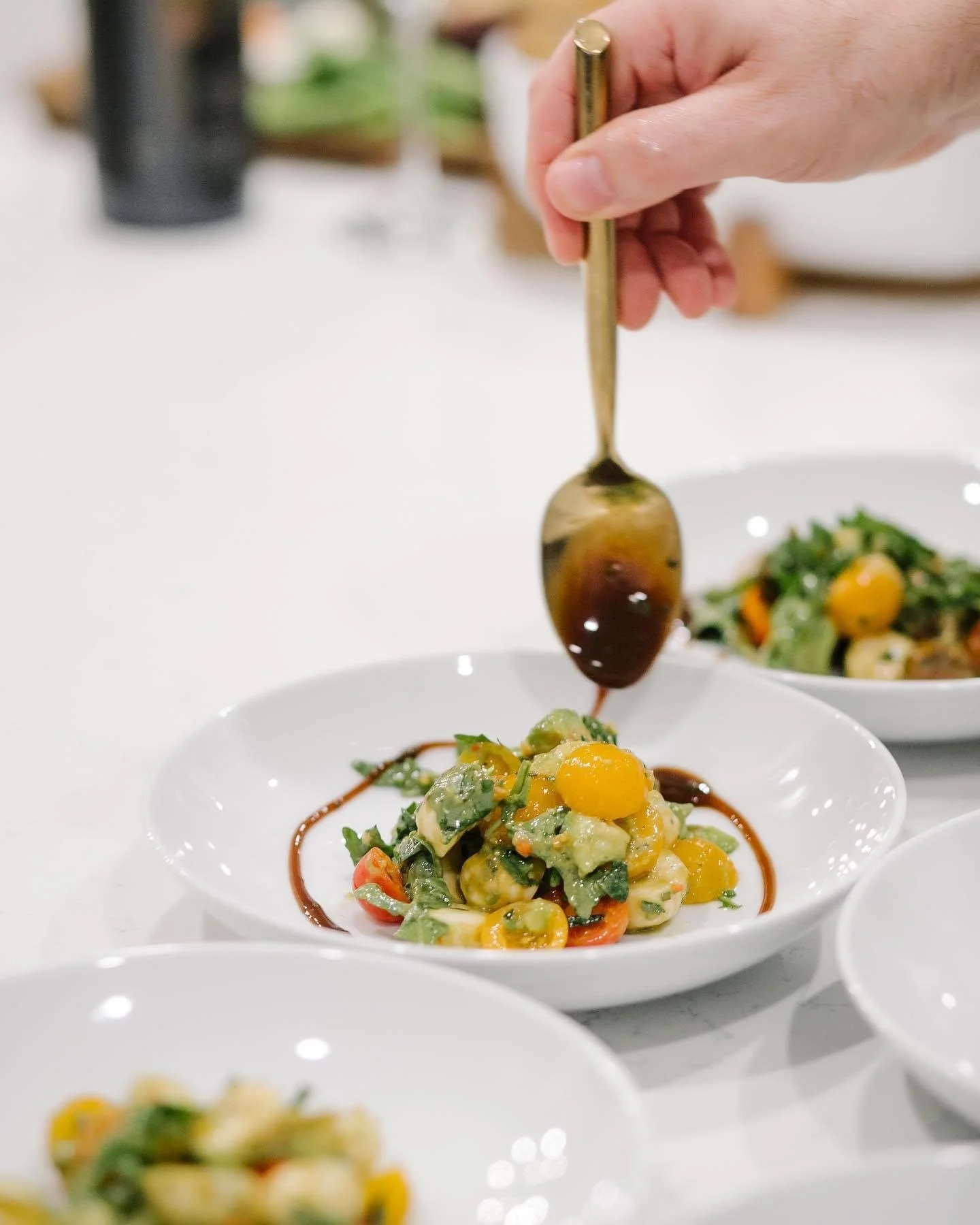 Person drizzling dark sauce over a colorful vegetable salad in a white bowl.