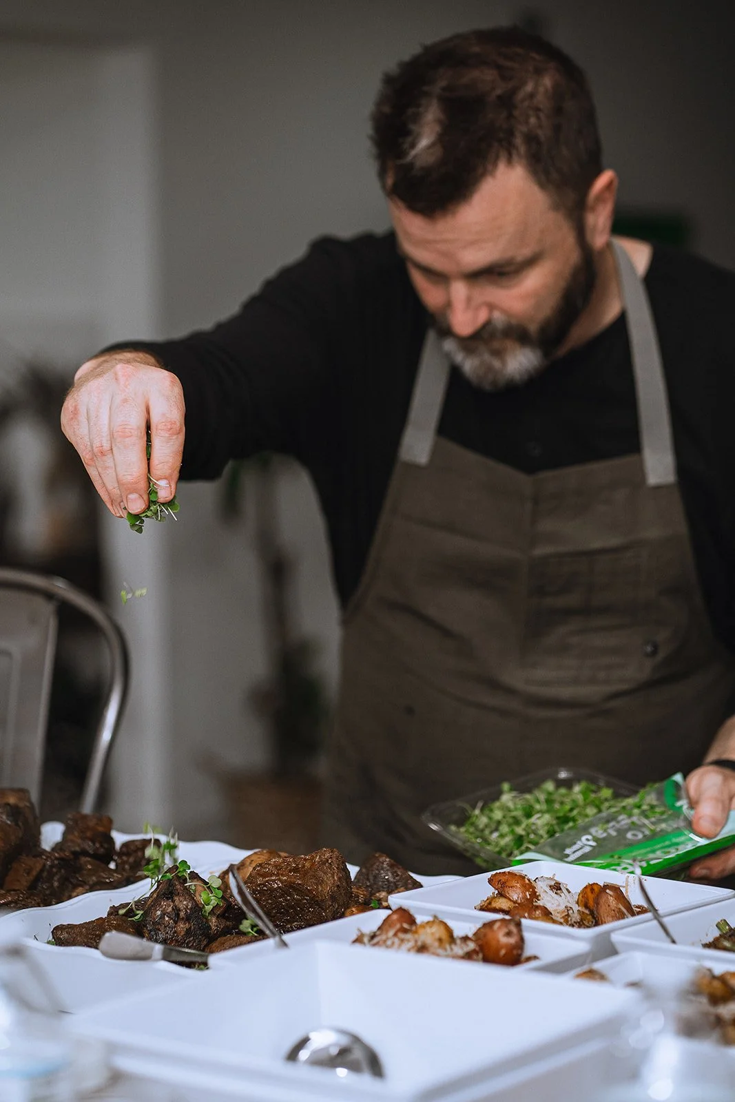 A man with dark hair and beard, wearing a black shirt and gray apron, is preparing food at a buffet-style setup with plates of cooked meat and greens.
