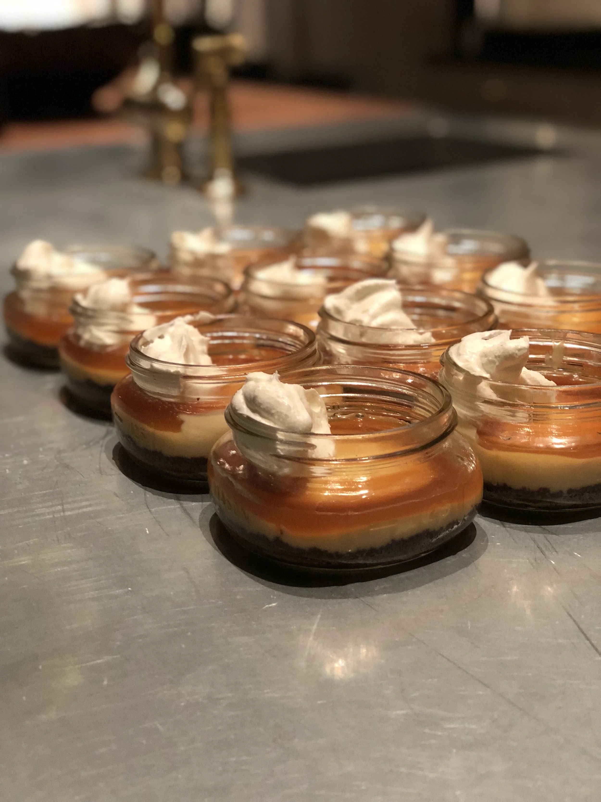 Small glass jars filled with layered dessert, topped with whipped cream, arranged in a row on a metallic surface.