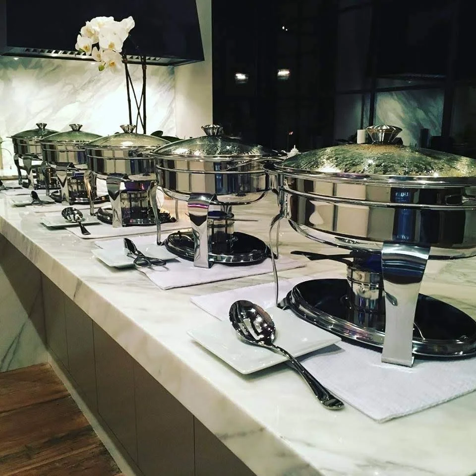 A row of six silver chafing dishes with lids, set on a white tablecloth with small white plates and spoons in front of each, in a restaurant or buffet setting.