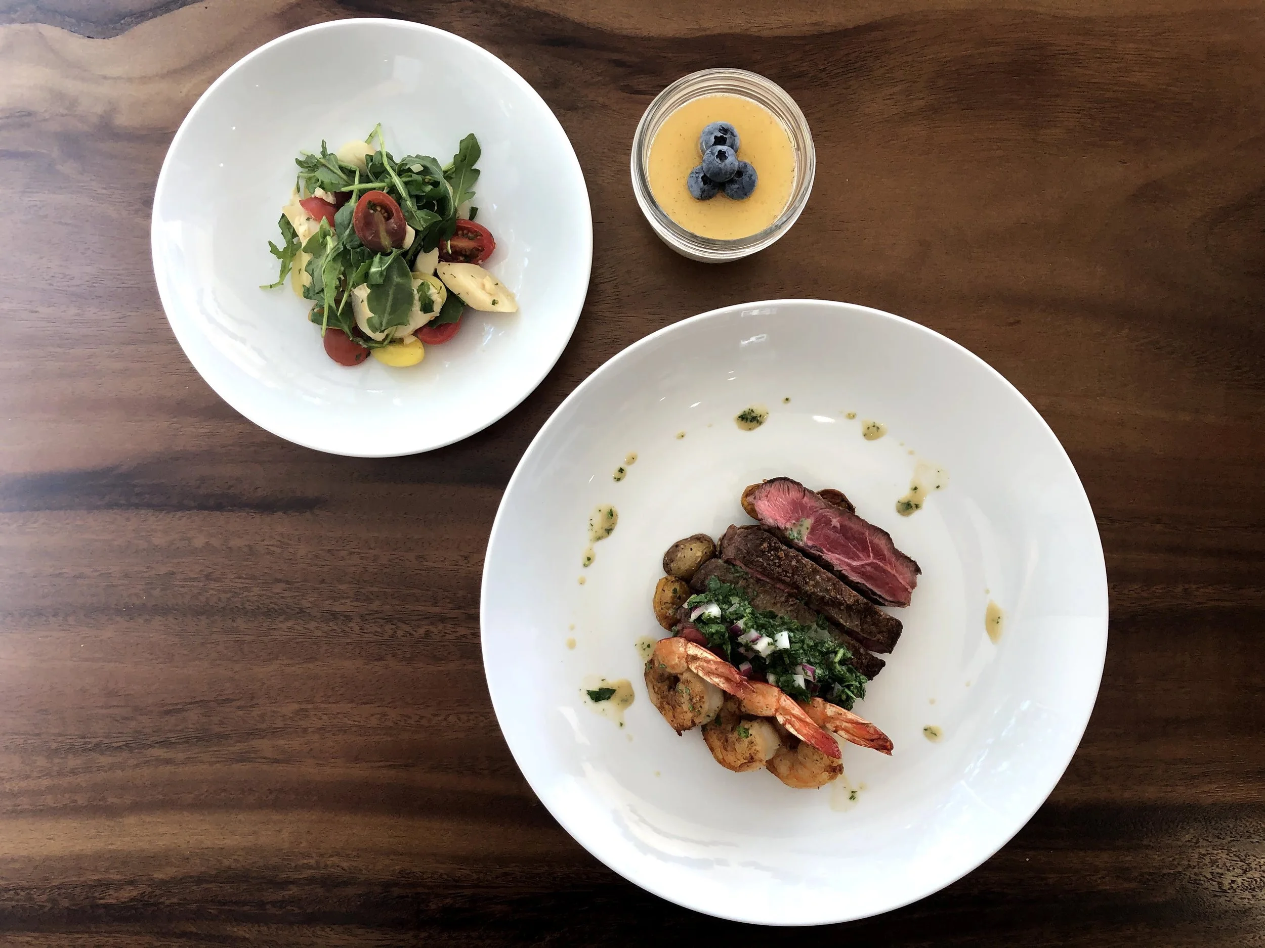 A white plate with slices of grilled steak, cooked shrimp, and topped with herbs, accompanied by a small side of roasted potatoes and sauce. Next to it, a smaller white bowl holds a colorful salad with arugula, cherry tomatoes, and cheese. A small gl