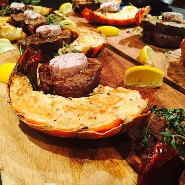 Lobster tail with baked stuffed beef medallions and lemon wedges on a wooden platter