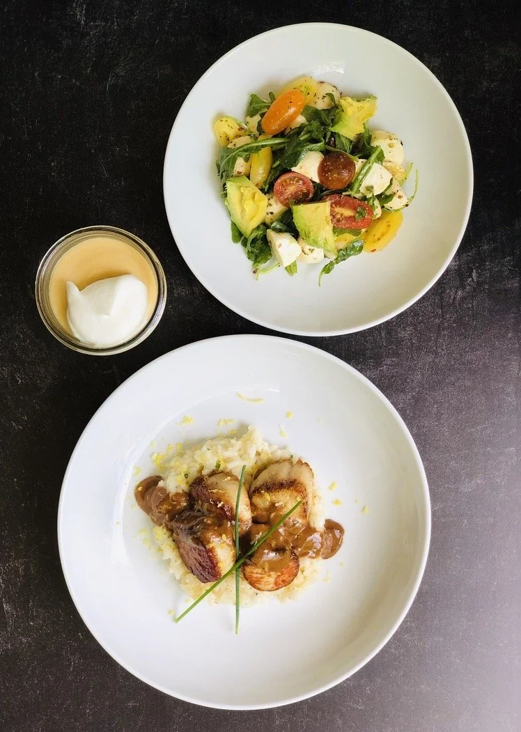 A meal with two white plates of food, a small glass of sauce or dressing with a dollop of white cream, and a dark table surface. The top plate contains a mixed salad with cherry tomatoes, avocado, mozzarella cheese, and greens. The bottom plate has a