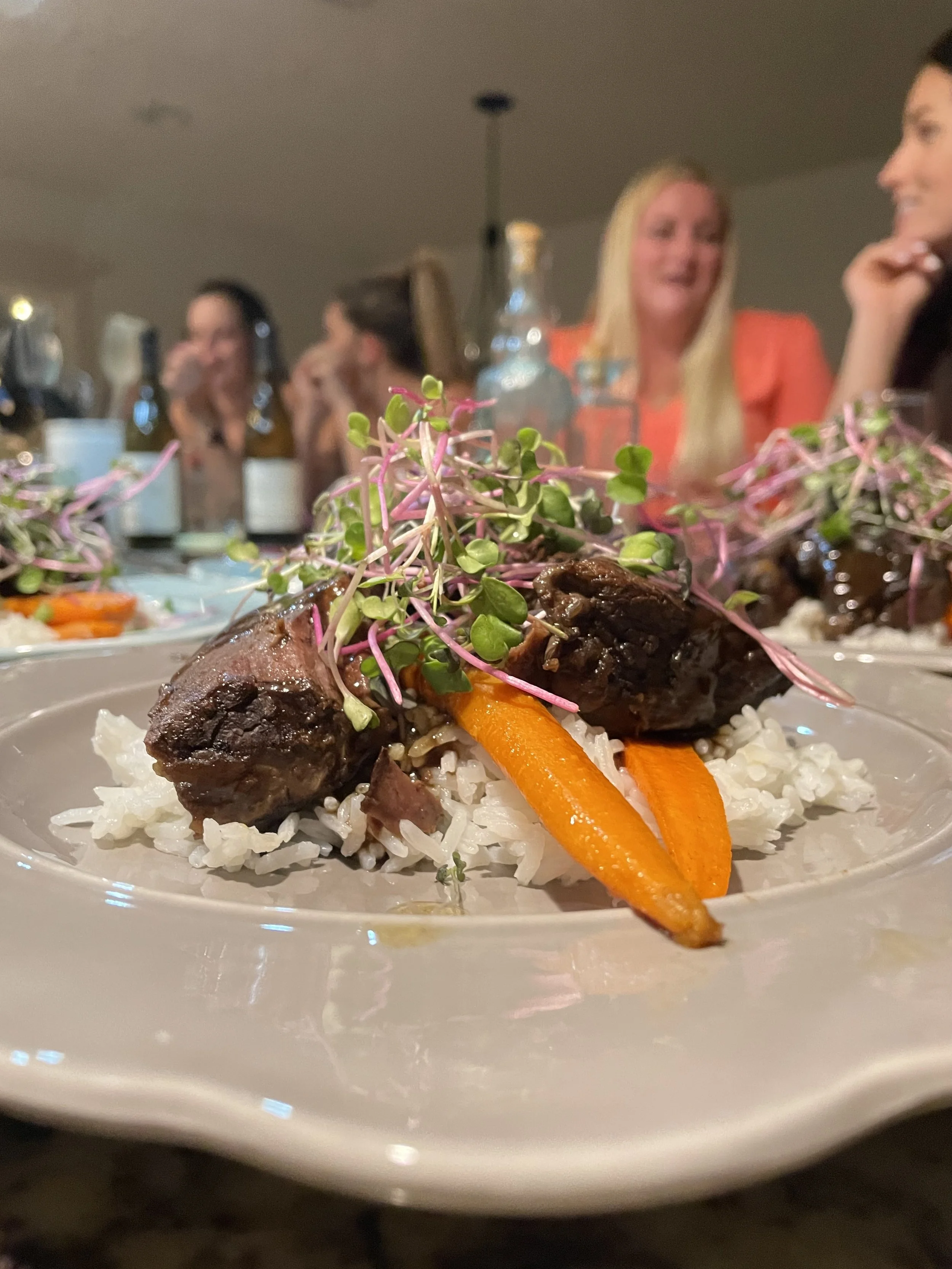 A plated serving of beef short ribs garnished with microgreens, served over rice with carrots in the foreground, at a dinner gathering with people dining and conversing in the background.