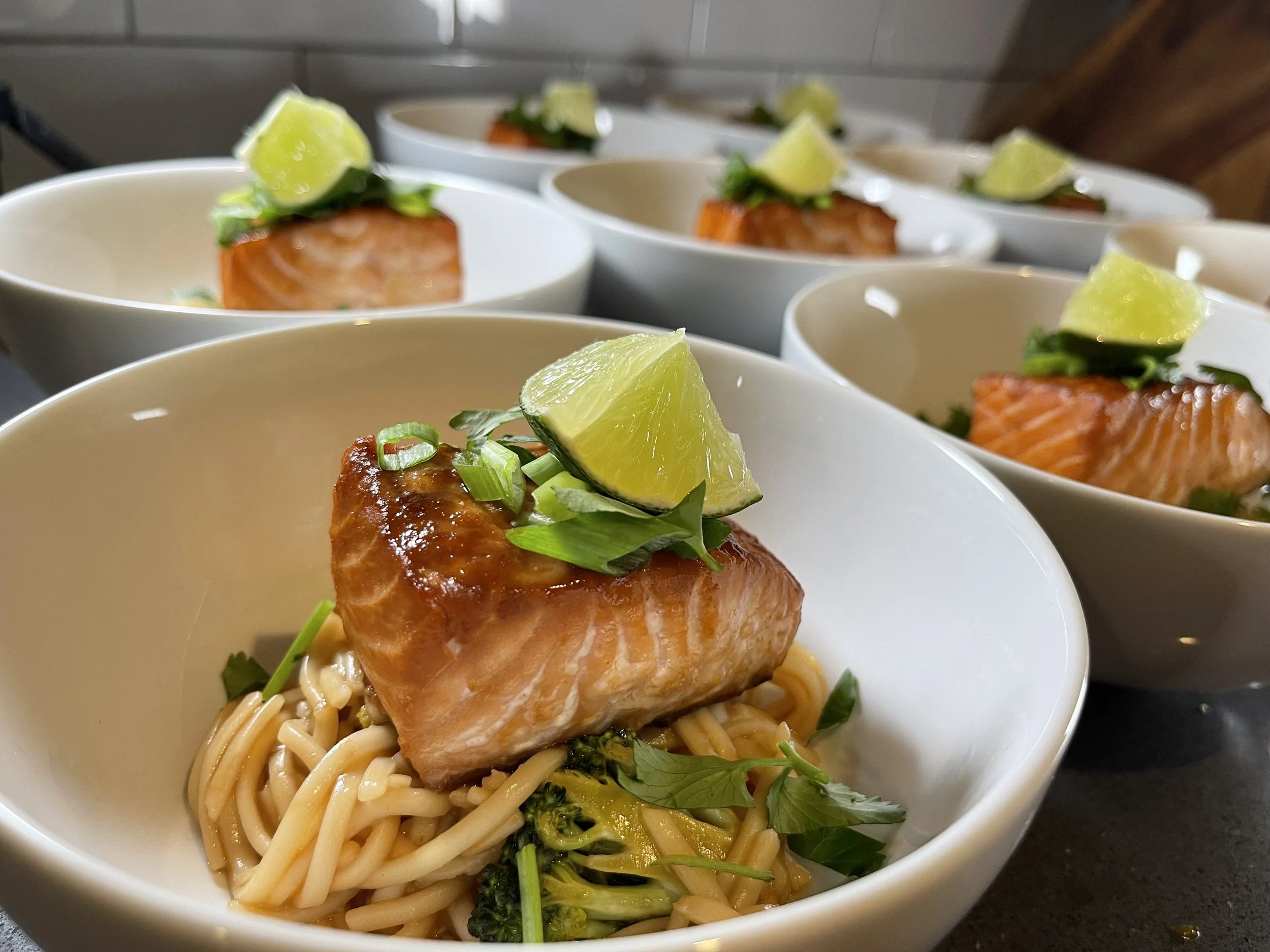 Bowl of cooked noodles topped with a cooked salmon piece, garnished with lime wedges, chopped green onions, and herbs, with similar bowls in the background.