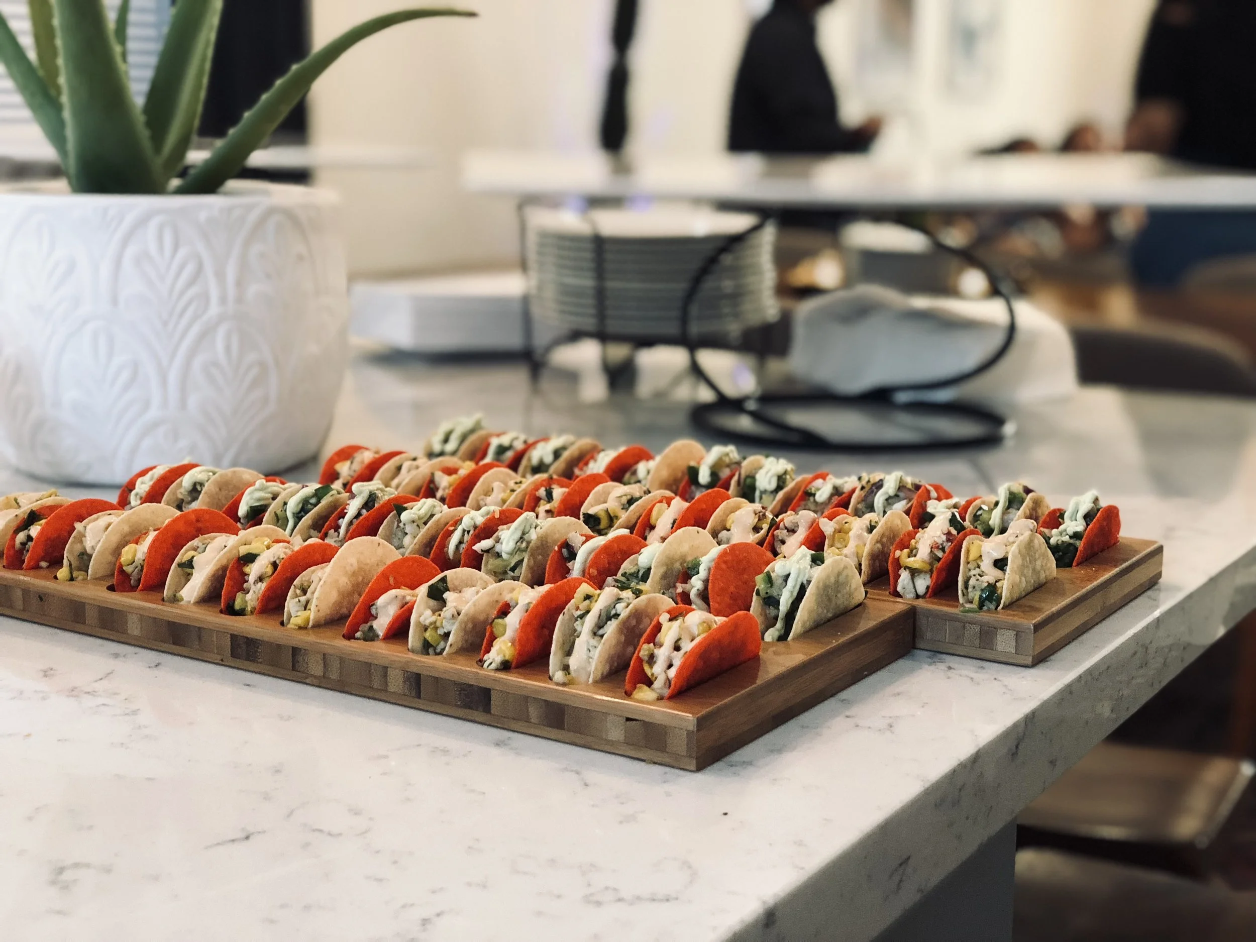 Tray of assorted mini tacos on a wooden board on a marble countertop, with a white potted plant and some electronic devices in the background.