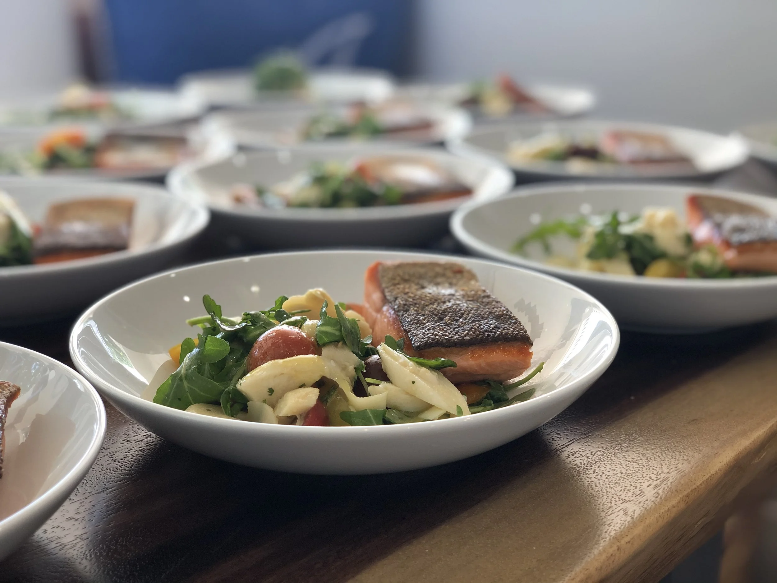 Close-up of a white bowl containing a side salad with greens, cherry tomatoes, and sliced cheese, accompanied by a piece of cooked salmon with crispy skin, placed on a dark wooden surface with blurred additional bowls of similar dishes in the backgro