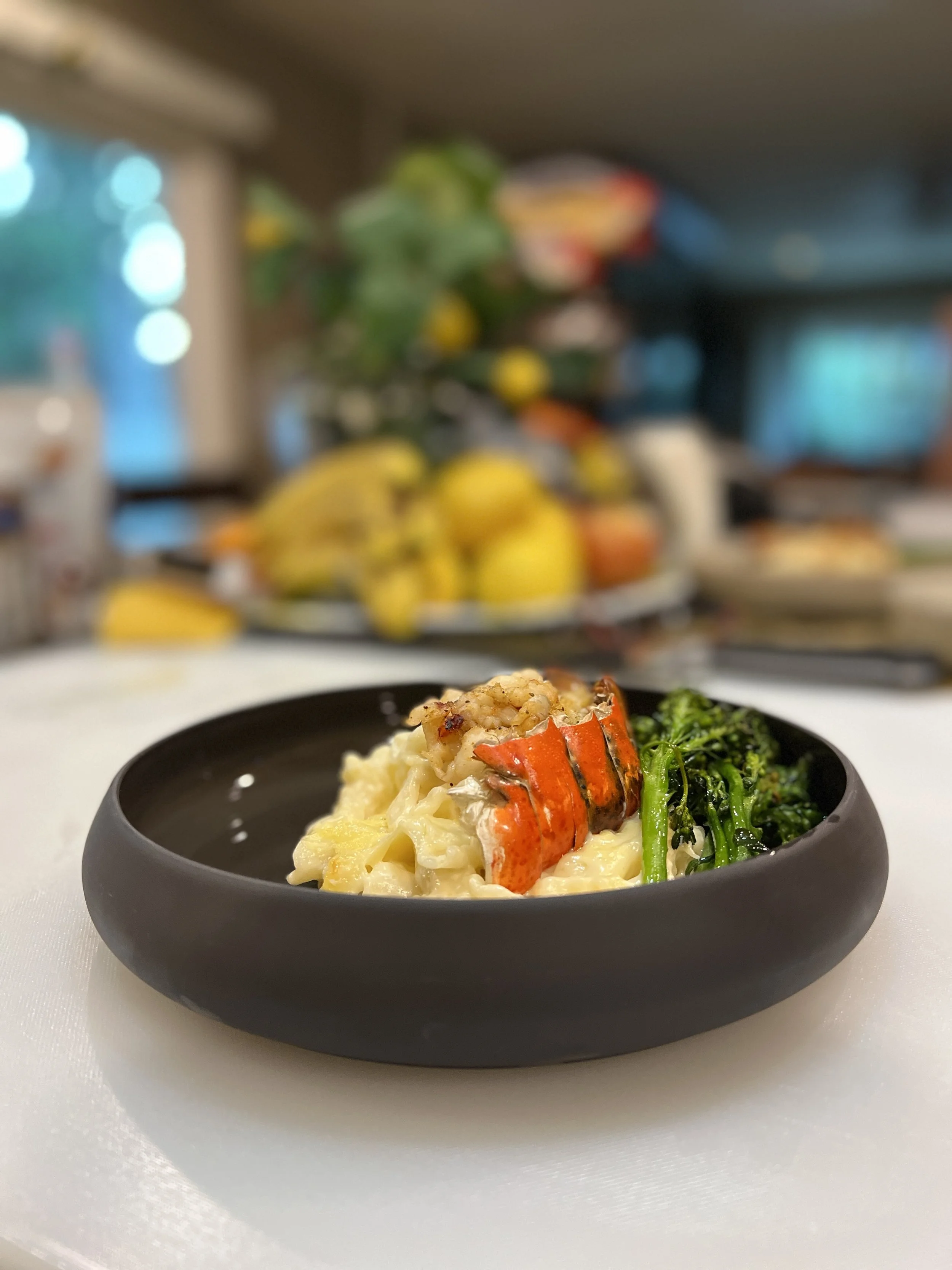 A black bowl containing macaroni and cheese with a piece of cooked lobster claw and a serving of greens on a white tablecloth with blurred kitchen background.