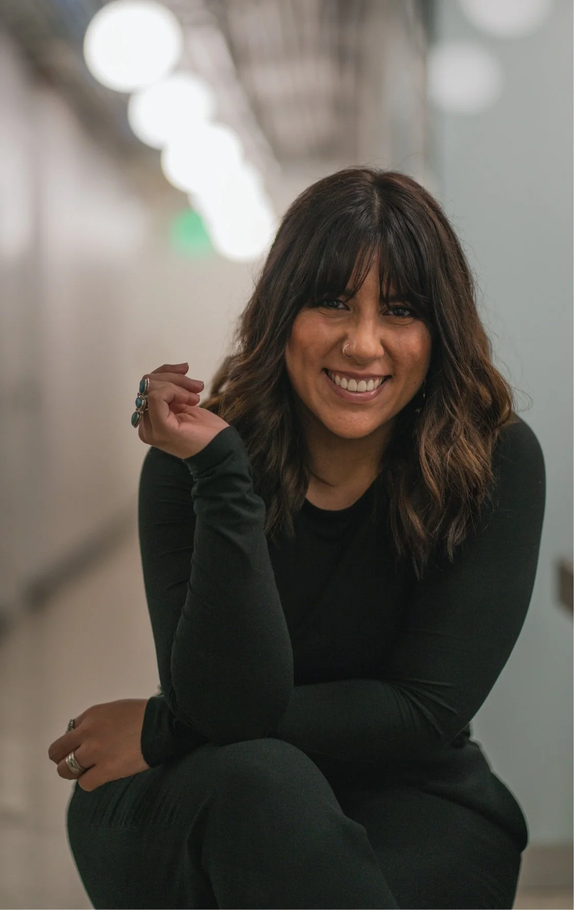 A photo of me smiling: Woman with wavy dark mid length hair, a nose ring, in a black long-sleeve top, sitting in a corridor with bright lights and blurred background.