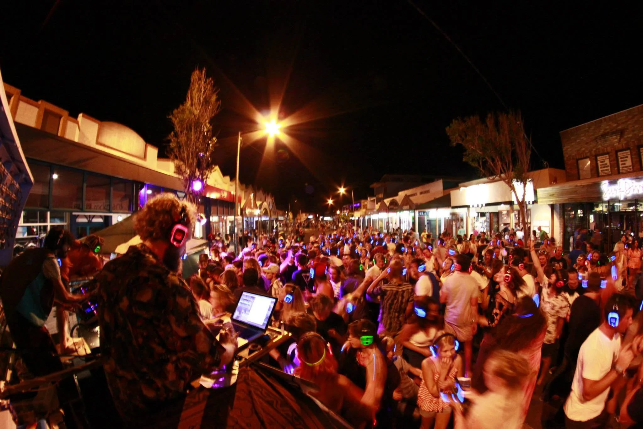 A crowded outdoor music event at night with people dancing and listening to a DJ on stage. Many are wearing colorful headphones, and the scene is lit with bright streetlights and colorful lighting at byron bay silent disco.