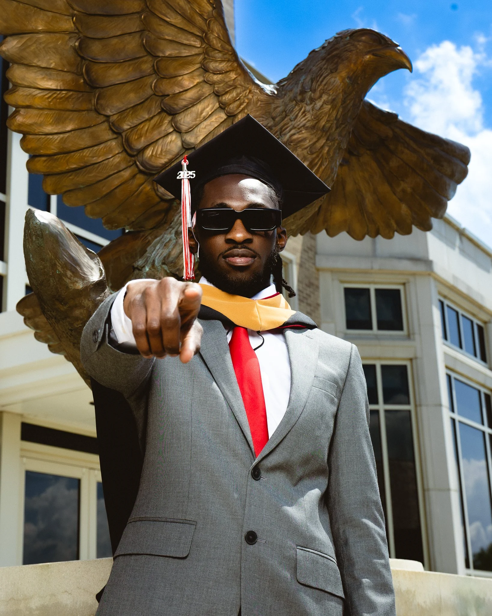 Graduating student in a gray suit, red tie, black sunglasses, black cap with "2025" tassel, standing in front of a large eagle statue, pointing towards the camera, on a campus with modern buildings under a blue sky.