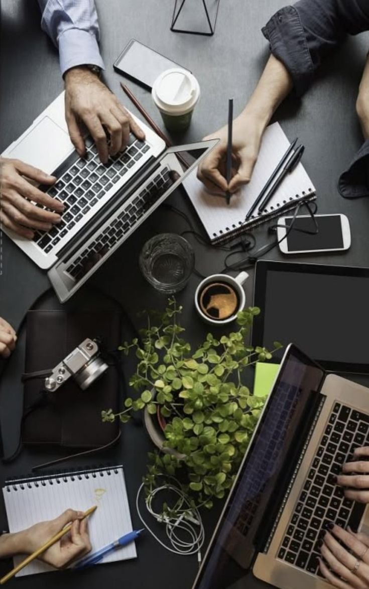 A workspace with multiple laptops, a smartphone, notebooks, pens, a camera, a cup of coffee, a glass of water, a plant, and people working at the table.